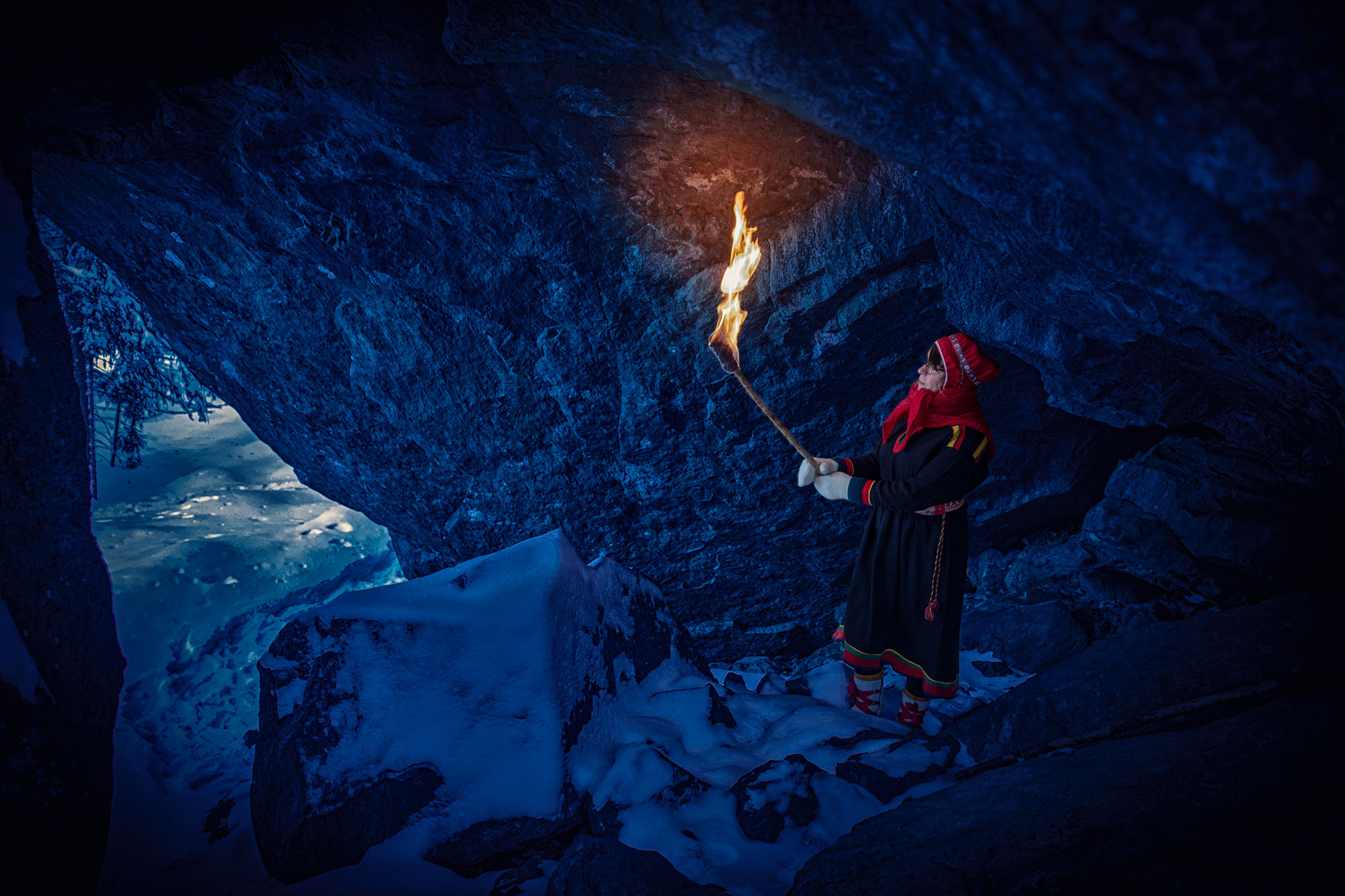 Inarin saamelainen perinneasussaan soihtu kädessä lumisessa Tynnyrikiven luolassa.